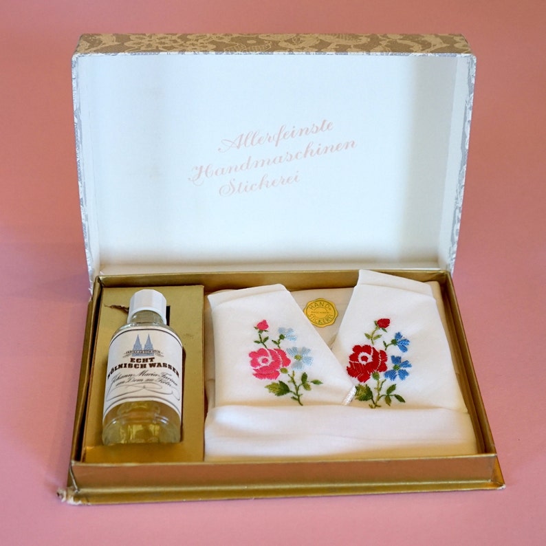 May include: A white box with a gold trim and a white lid with the text "Allerfeinste Handmaschinenstickerei" in pink lettering. The box contains two white handkerchiefs with embroidered flowers and a small clear bottle with a label that reads "Echt Almrich Wasser".