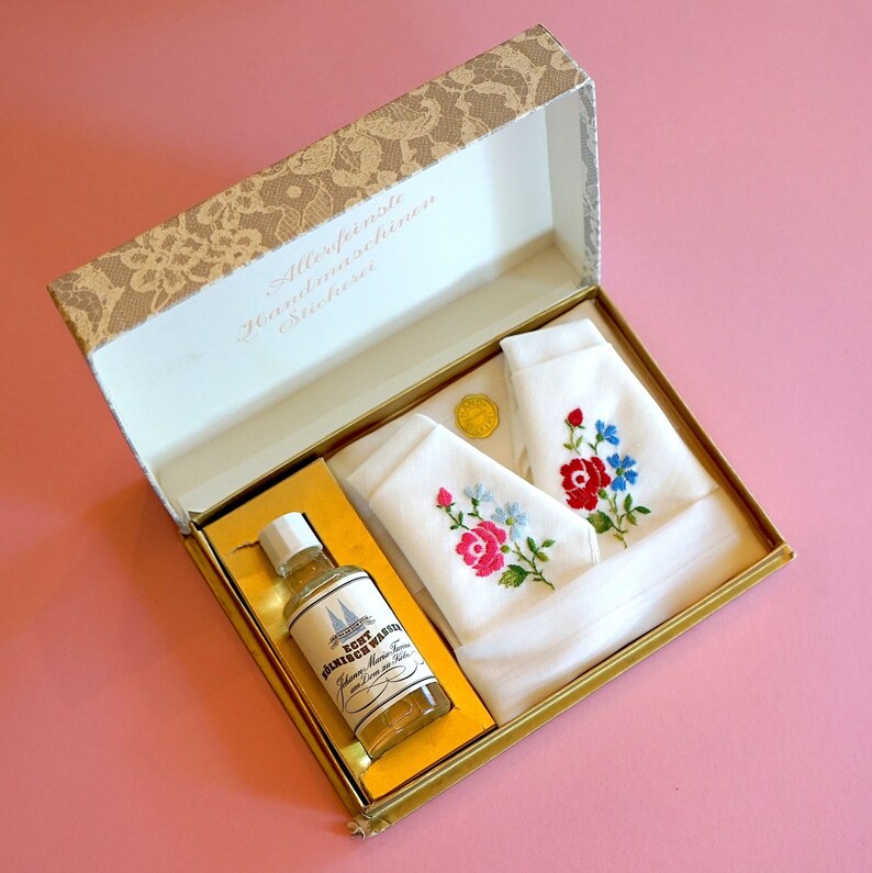 May include: A white gift box with a gold trim and a lace design on the lid. The box contains two white handkerchiefs with embroidered flowers and a small bottle of clear liquid with a label that reads "Fein Waschmittel".