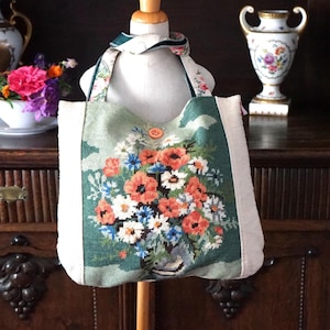 Könnte beinhalten: Eine Gobelin-Tragetasche mit einem Blumendesign mit orangefarbenen Mohnblumen, weißen Gänseblümchen und blauen Blumen auf grünem Hintergrund. Die Tasche hat einen cremefarbenen Rand und Träger sowie einen Holzknopfverschluss. Die Tasche wird auf einer Schaufensterpuppe präsentiert.