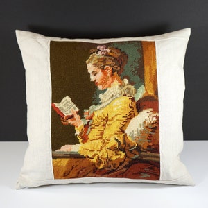 Può includere: Un cuscino color crema con un'immagine ricamata quadrata di una donna in un vestito giallo che legge un libro. Il ricamo è in vari colori, tra cui rosso, verde e marrone.