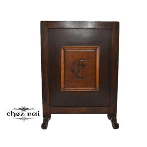 May include: A dark brown wooden fireplace screen with a carved wooden panel featuring the letter "C". The screen has three legs.