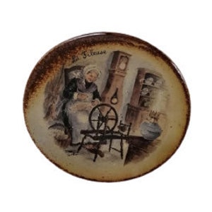 May include: A ceramic plate with a brown and beige background featuring a woman spinning a wheel in a room with a clock and a fireplace. The text "La Fileuse" is written on the plate.