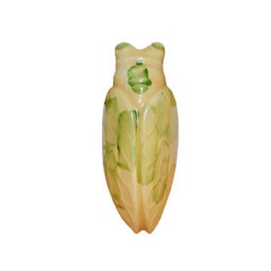 May include: A light yellow ceramic figurine of a cicada with green painted details. The figurine is shaped like a cicada with wings and a long abdomen.