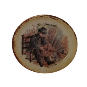 May include: A round, cream-colored ceramic plate with a painted image of a man sitting on a chair, weaving a basket. The plate has the text "Le Vannier" in brown lettering.