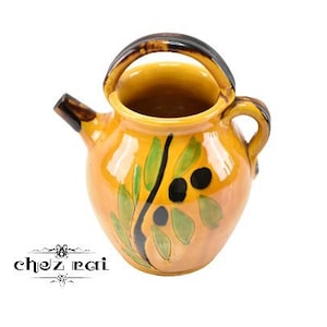 Puede incluir: Una jarra de cerámica amarilla con un asa negra y un diseño de rama de olivo. La jarra tiene una boca ancha y un cuerpo redondeado.