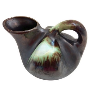 Peut inclure: Un petit pichet en céramique brun avec un glaçage vert et brun. Le pichet a un corps arrondi et une anse.