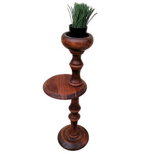 May include: A dark brown wooden plant stand with a small round shelf and a top section holding a black pot with green grass. The stand has a turned design and a round base.