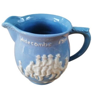 May include: A light blue ceramic pitcher with a white design of people riding on a camel. The pitcher has a handle and says "Widecombe Fair" on the side.