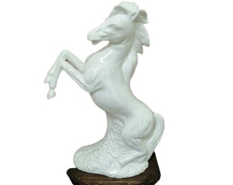 Puede incluir: Estatua de caballo de cerámica blanca con una pata delantera levantada, de pie sobre una base. El caballo está en una pose de alzamiento.
