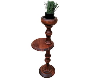 May include: A tall, dark brown wooden plant stand with a small round shelf and a top section holding a black pot with green grass. The stand has a turned design and a round base.