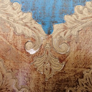 May include: Close-up of a wooden surface with a gold leaf design. The design features intricate swirls and leaves, creating a decorative and elegant pattern. The surface is distressed, adding to its vintage charm.