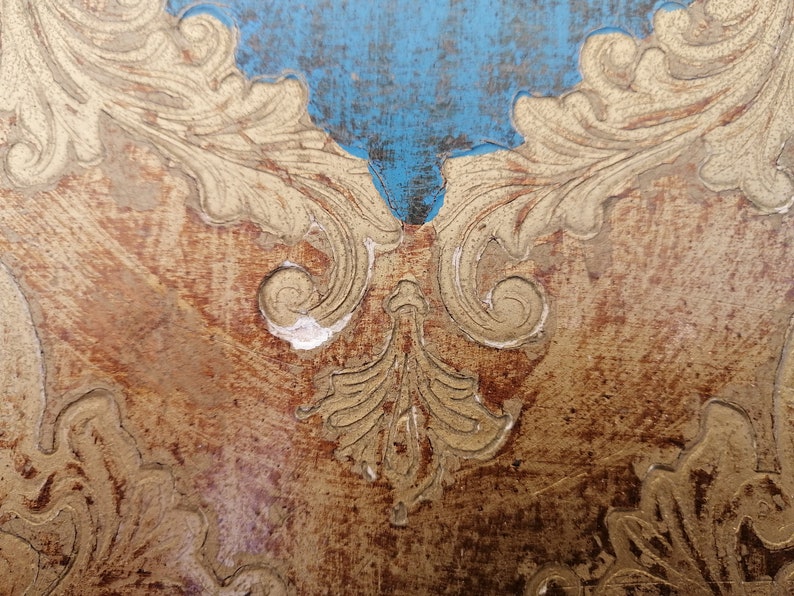 May include: Close-up of a wooden surface with a gold leaf design. The design features intricate swirls and leaves, creating a decorative and elegant pattern. The surface is distressed, adding to its vintage charm.