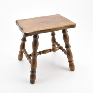 Puede incluir: Un pequeño taburete de madera con una tapa rectangular y patas torneadas. El taburete está hecho de madera marrón oscura y tiene un aspecto rústico.