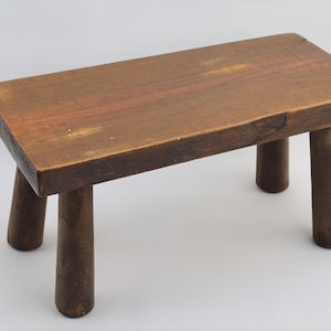 May include: A rustic wooden stool with a rectangular top and four turned legs. The stool is made of dark brown wood and has a natural finish.