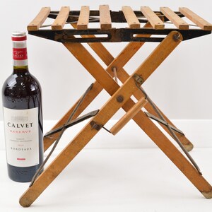 May include: A wooden folding stool with a black metal frame and a slatted top. The stool is in a folded position and is standing on a white surface. A bottle of wine is in the background.