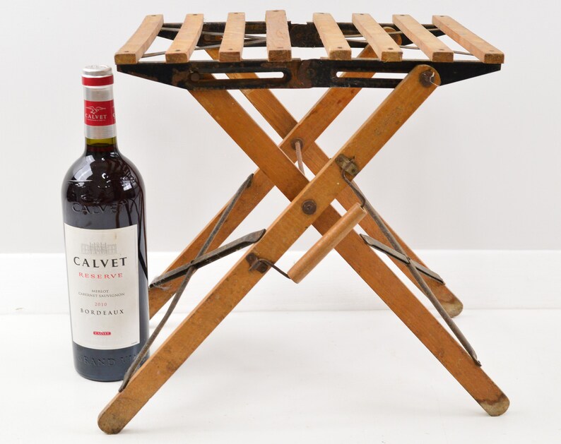 May include: A wooden folding stool with a black metal frame and a slatted top. The stool is in a folded position and is standing on a white surface. A bottle of wine is in the background.