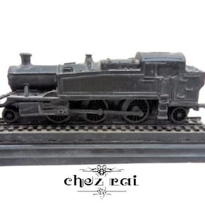 May include: A dark gray model train on a track base. The train features a detailed engine, smokestack, and multiple wheels. The base has the text "chez rai" in a decorative font.