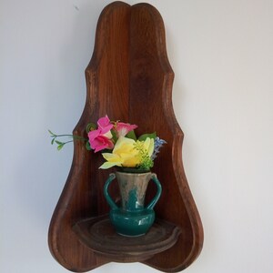 May include: A wooden corner shelf with a carved design, featuring a green ceramic vase with pink and yellow flowers.