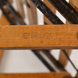May include: Close-up of a wooden folding chair with a black metal frame. The chair has a wooden seat and back, and the frame is made of metal rods. The chair is stamped with the word "EUREKA" on the underside of the seat.