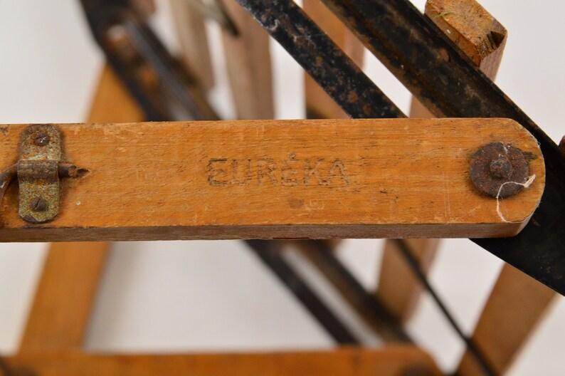 May include: Close-up of a wooden folding chair with a black metal frame. The chair has a wooden seat and back, and the frame is made of metal rods. The chair is stamped with the word "EUREKA" on the underside of the seat.
