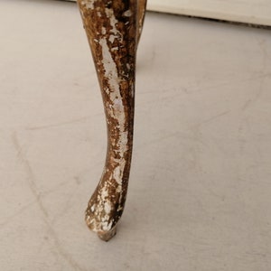 May include: Close-up of a wooden table leg with a distressed white paint finish. The leg is carved with a decorative design and has a slightly curved shape.