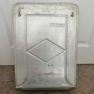 Puede incluir: Un letrero de metal plateado con una forma de diamante en el centro. El letrero tiene dos agujeros en la parte superior para colgarlo.