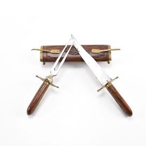 Może przedstawiać: Drewniana pochwa z mosiężnym zatrzaskiem zawiera srebrny nóż i widelec z drewnianymi uchwytami. Nóż i widelec są skrzyżowane wewnątrz pochwy.