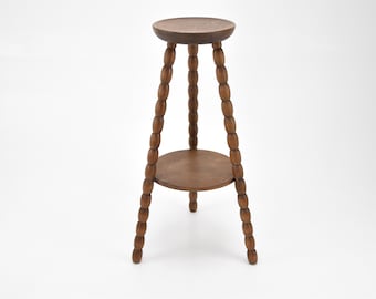 May include: A two-tiered wooden plant stand with a brown finish. The stand has three legs with a beaded design.