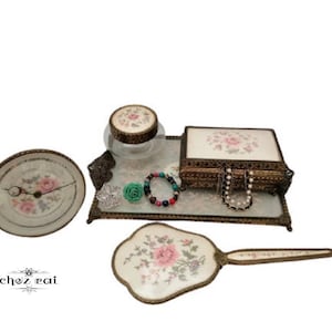May include: A vintage vanity set with a glass jar, a mirrored tray, a small round mirror, a hand mirror, and a jewelry box. The set features floral designs and gold accents.