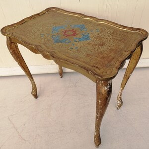 May include: A gold-painted side table with a decorative, ornate top and three legs. The table has a distressed finish, giving it a vintage look.