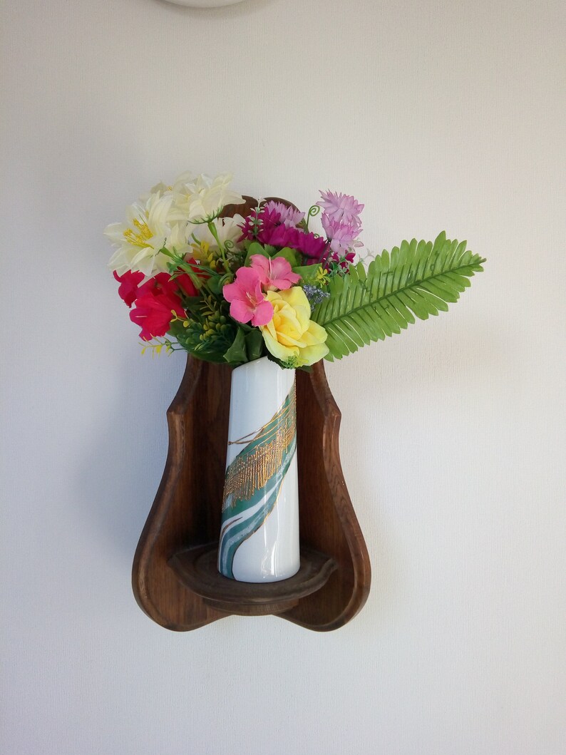 May include: A wooden shelf shaped like a violin with a white vase with green and gold designs holding a bouquet of pink, yellow, white, and red flowers.