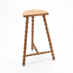 Könnte beinhalten: Ein hellbrauner Holzstuhl mit halbmondförmigem Sitz. Der Hocker hat drei Beine mit einem gedrehten, seilartigen Design. Die Beine sind durch eine horizontale Strebe nahe der Basis verbunden. Der Hocker ist aus Holz gefertigt und hat eine natürliche Oberfläche.