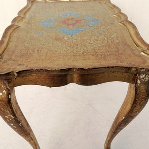 May include: A gold-painted side table with a scalloped top and intricate floral design. The table has three legs with a curved design.