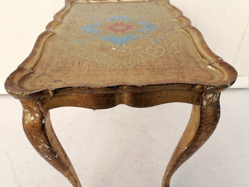 May include: A gold-painted side table with a scalloped top and intricate floral design. The table has three legs with a curved design.