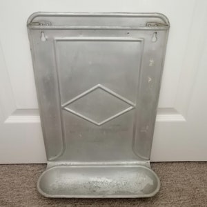 Puede incluir: Una panera de pared vintage de metal con un patr&oacute;n de diamante y una bandeja extra&iacute;ble. La panera es de color plateado y tiene dos agujeros para colgar.