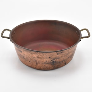 May include: A round copper cooking pot with two riveted metal handles on opposite sides. The exterior surface shows a weathered, scratched patina with dark marks, while the interior is smooth and reddish-brown in colour with a wide opening.