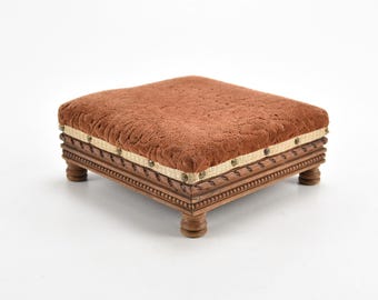 May include: A brown upholstered footstool with a wooden frame and four turned legs. The top of the footstool is covered in a brown fabric with a woven pattern. The frame is decorated with a carved design and has a beige trim.