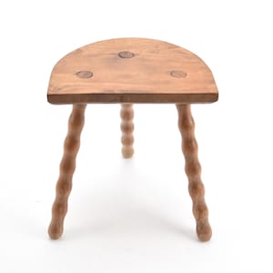 May include: A small, three-legged wooden stool with a half-moon shaped seat. The legs are carved with a spiral design. The stool is made of light-colored wood and has a natural finish. The stool is approximately 12 inches tall.