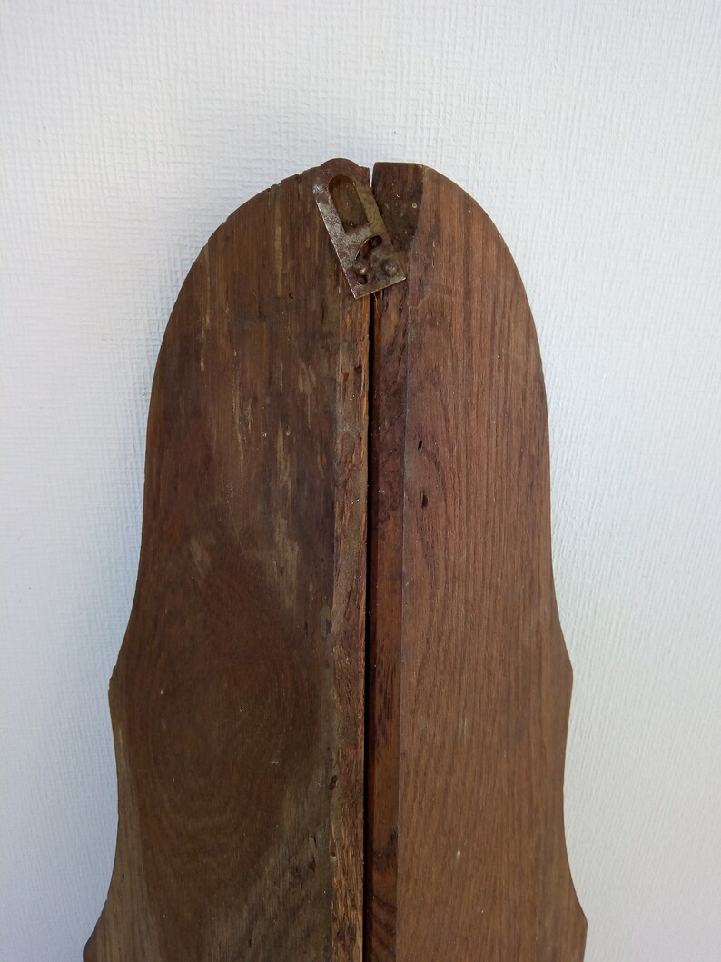 May include: The back of a wooden frame with a decorative, scalloped edge. The frame is made of two pieces of wood that are joined together with a metal hinge. The wood is a dark brown colour and has a natural wood grain.