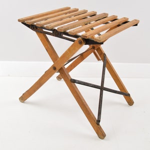 May include: A wooden folding stool with a slatted seat and black metal supports. The stool is made of light brown wood and has a simple, functional design.