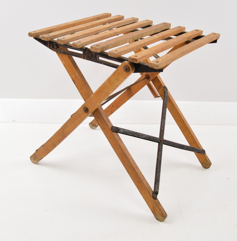 May include: A wooden folding stool with a slatted seat and black metal supports. The stool is made of light brown wood and has a simple, functional design.