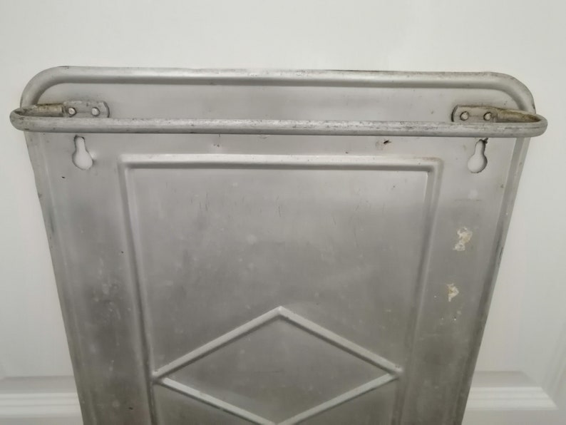 Puede incluir: Un estante de pared de metal plateado con un patr&oacute;n de diamante. El estante tiene dos agujeros en la parte superior y dos agujeros en la parte inferior para colgar.