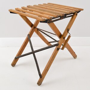 May include: A wooden folding stool with a slatted seat and a metal frame. The stool is in a folded position and is ready to be used.