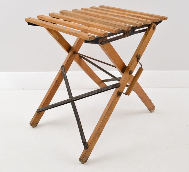 May include: A wooden folding stool with a slatted seat and a metal frame. The stool is in a folded position and is ready to be used.