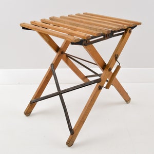 May include: A wooden folding stool with a slatted seat and metal supports. The stool is made of natural wood and has a rustic look.