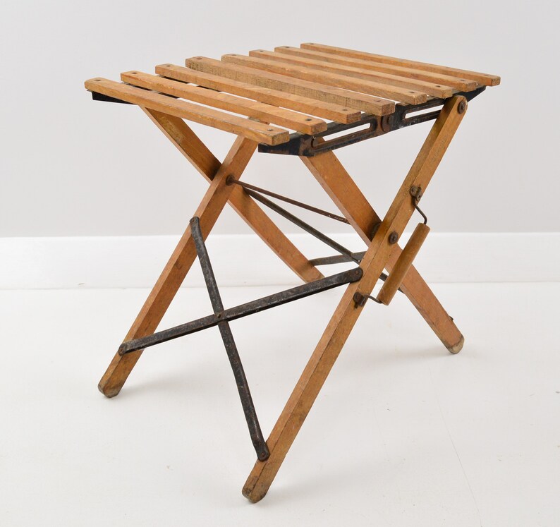 May include: A wooden folding stool with a slatted seat and metal supports. The stool is made of natural wood and has a rustic look.