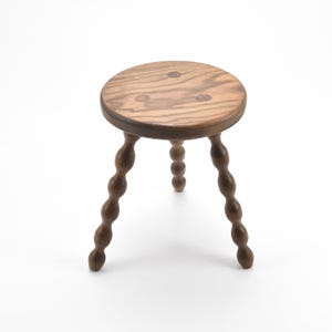 May include: A small, wooden stool with a round seat and three turned legs. The seat is light brown with visible wood grain, and the legs are a darker brown. The legs have a distinctive, bulbous design. The stool is set against a white background.