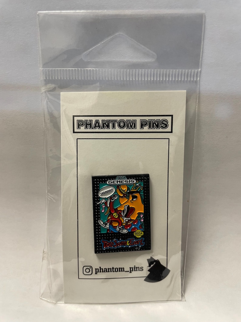 May include: A pin featuring a colourful graphic from the Sega Genesis game, ToeJam & Earl. The pin is rectangular with a black border and the game's logo. The pin is on a white card with "Phantom Pins" printed on it.