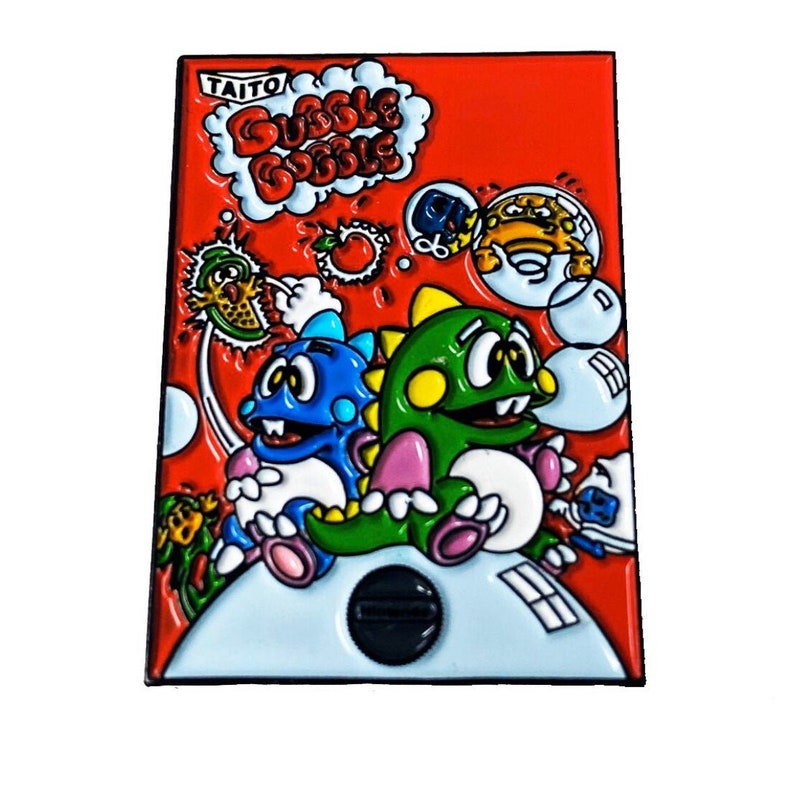 May include: A red enamel pin featuring the characters from the classic arcade game Bubble Bobble. The pin shows the two main characters, a blue and green dinosaur, with the game's title in a thought bubble above them.