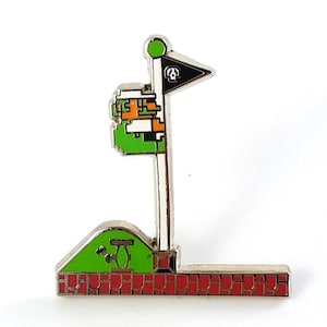 May include: A silver-colored pin featuring a pixelated golf flag design. The flag is black with a white skull and crossbones. The pin includes a green hill, red brick base, and a pixelated character. The flag pole has a green circle at the top.
